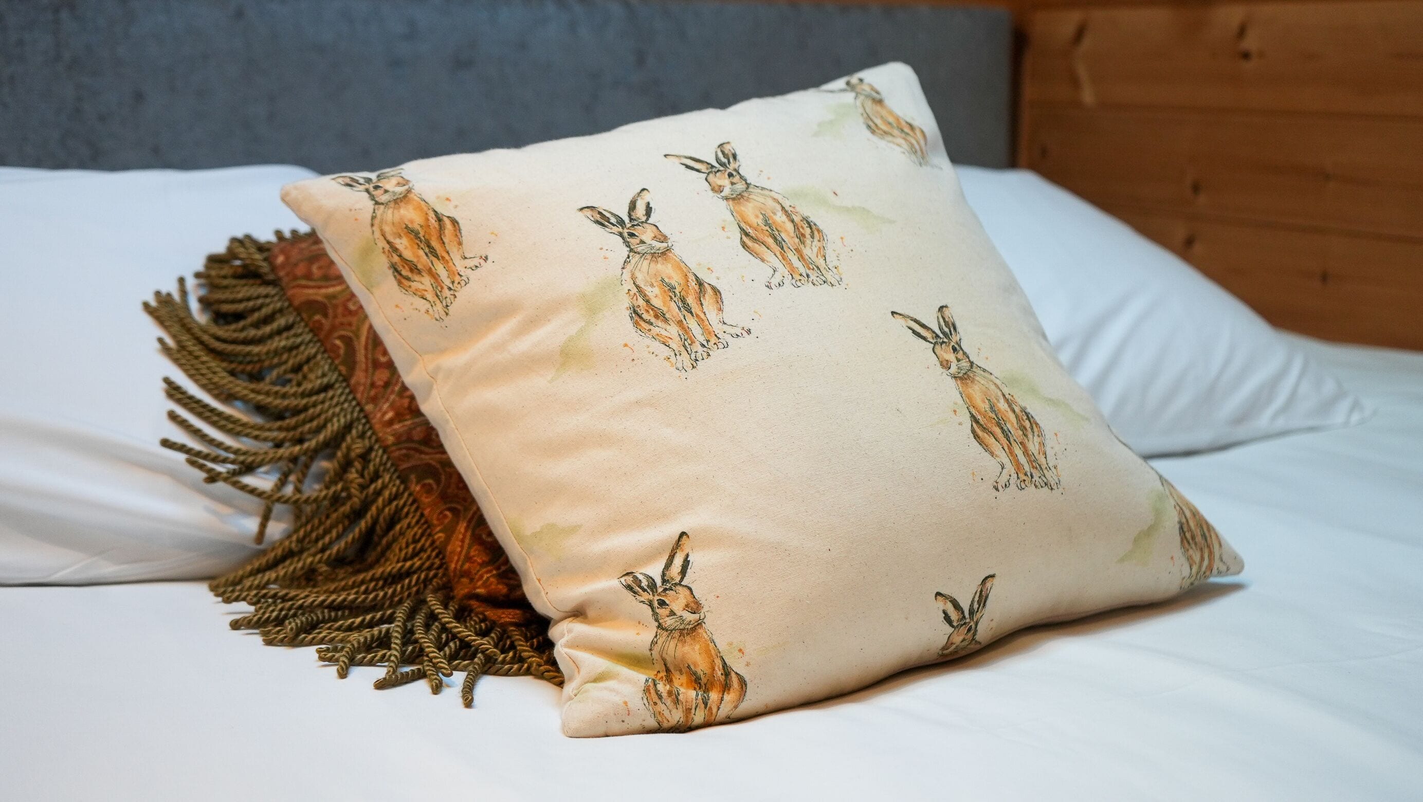 Un lit avec un drap blanc, un oreiller beige orné d’illustrations de lapins et un plaid marron tissé.