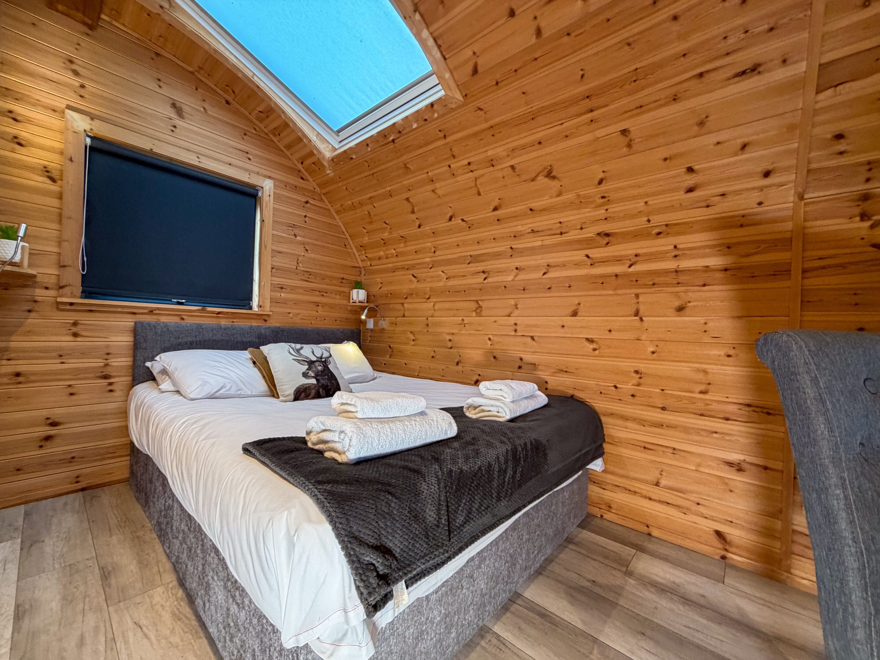 Une chambre confortable dans un chalet en bois, avec une lucarne, un lit orné d’un coussin à motif de cerf et une table de chevet avec une lampe.