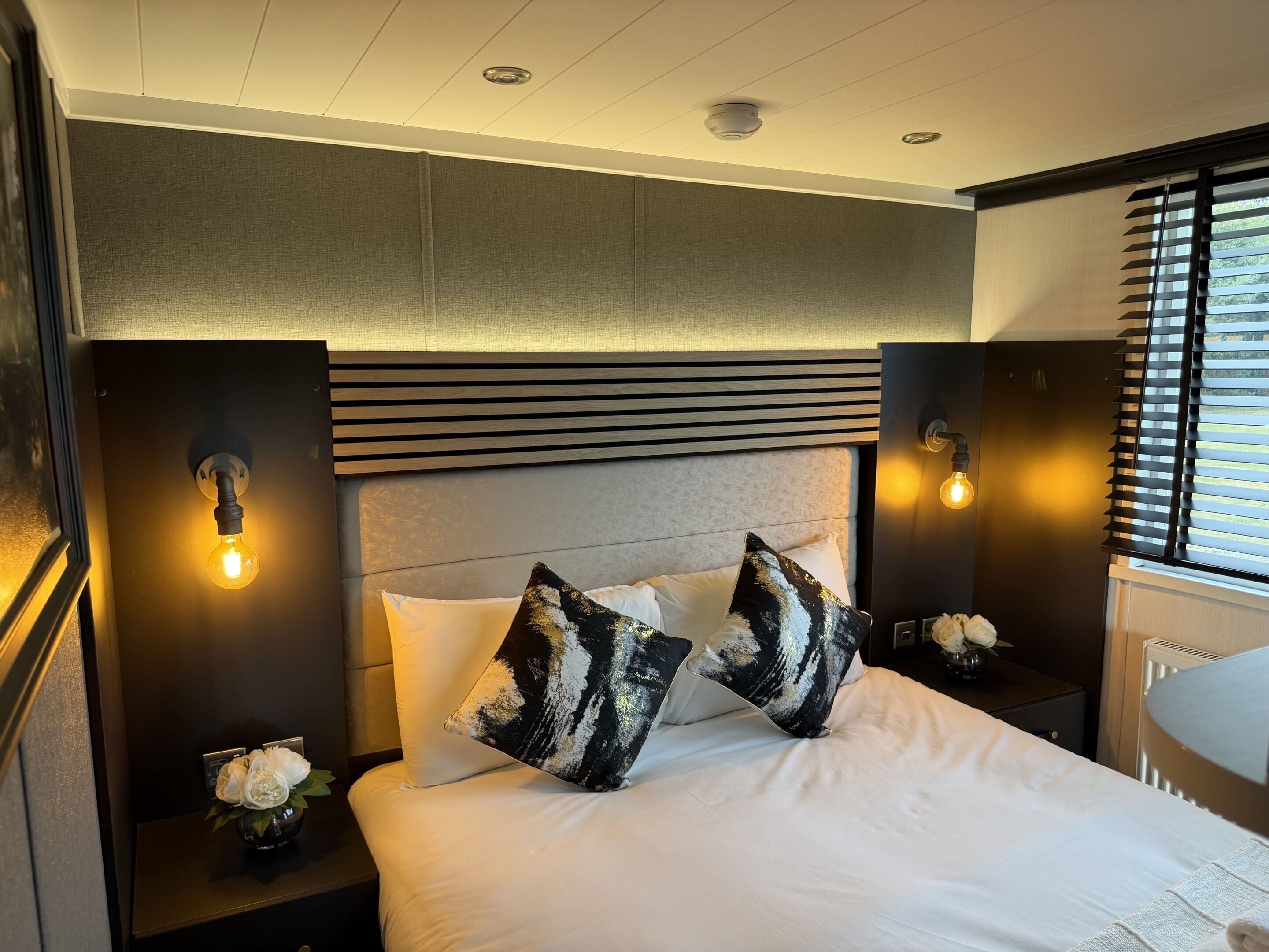 Habitación de hotel con cama, dos mesitas de noche, una lámpara de pared y una ventana con persianas.