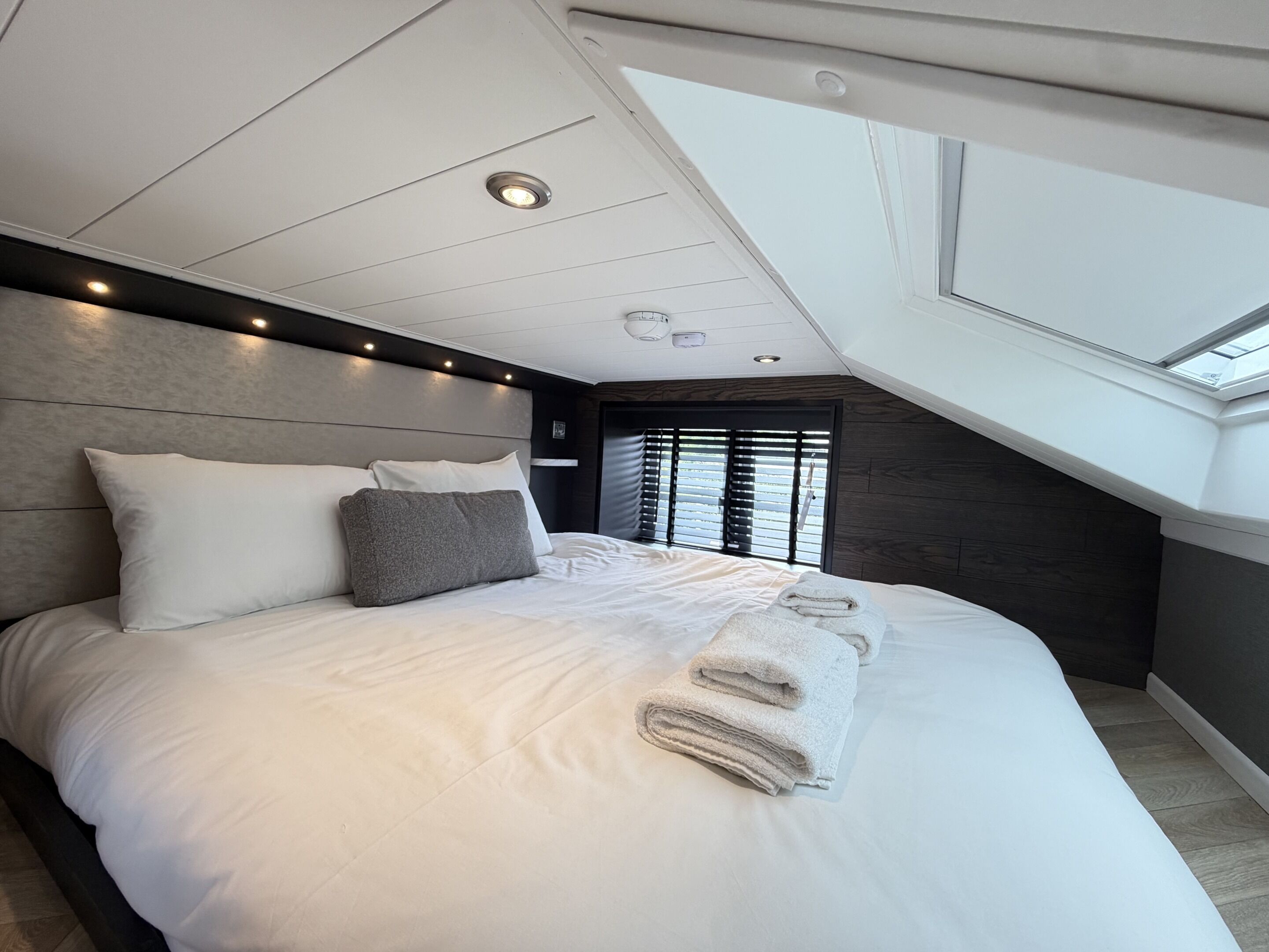 Une chambre à coucher moderne avec une lucarne, un grand lit recouvert de draps blancs et une tête de lit sombre.