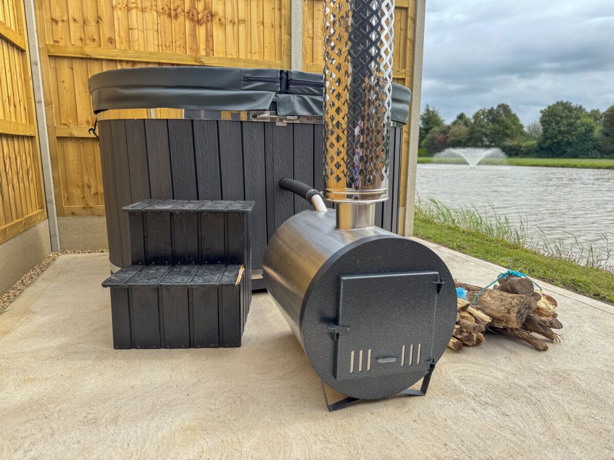 A wood stove with a chimney, a bench, and logs on a concrete surface near a wooden fence and a body of water.