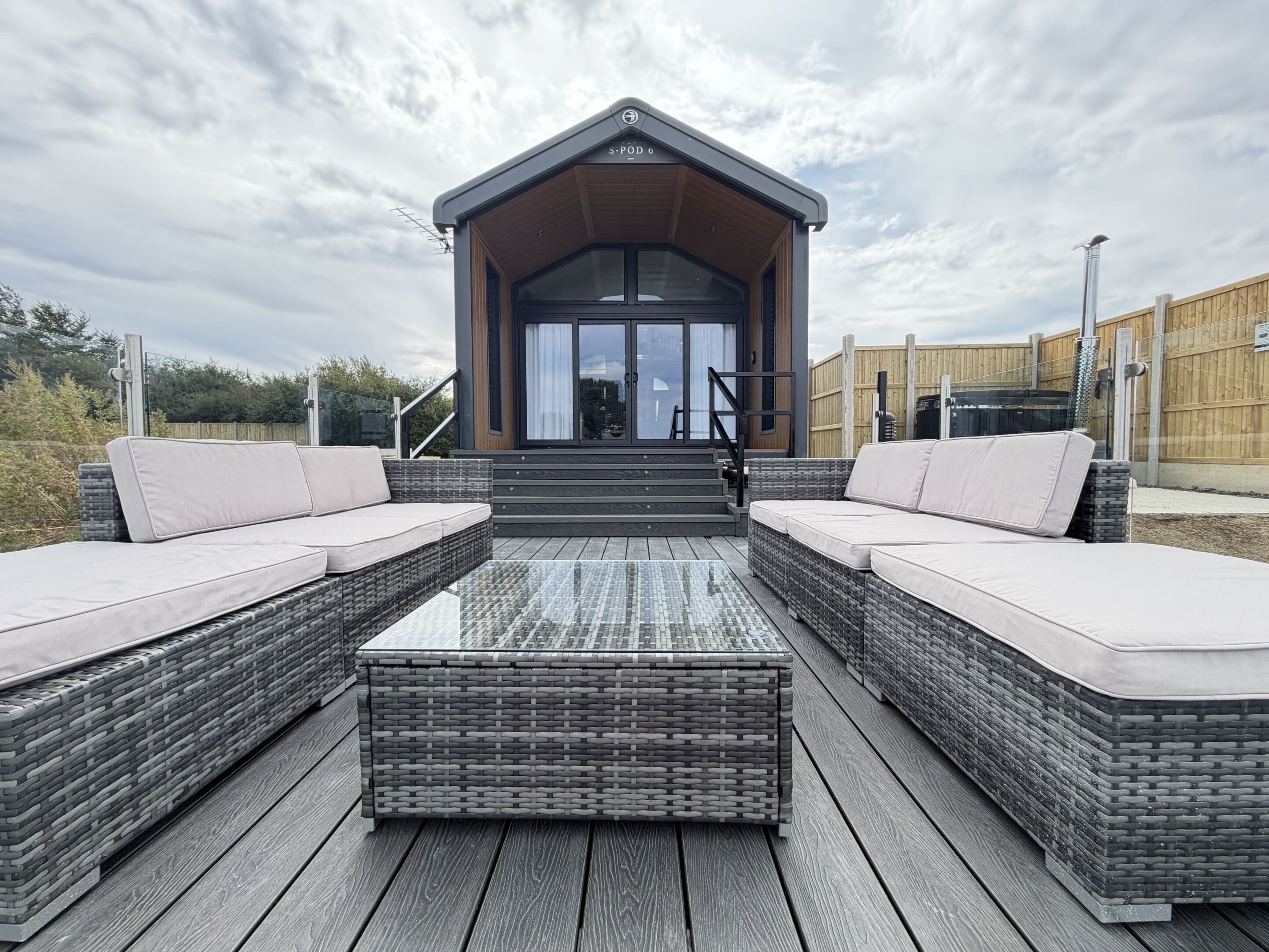 A modern outdoor seating area with wicker furniture and a glass table, leading to a building with a sloped roof and a sign reading 'S-POD 6'.