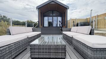 A modern outdoor seating area with wicker furniture and a glass table, leading to a building with a sloped roof and a sign reading 'S-POD 6'.
