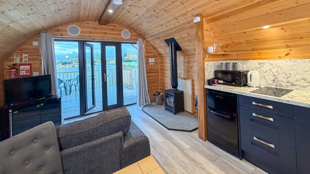 Interior acogedor de una cabaña con techo de madera, sofá, zona de cocina con electrodomésticos modernos y puerta de vidrio corredera que da a una terraza exterior.