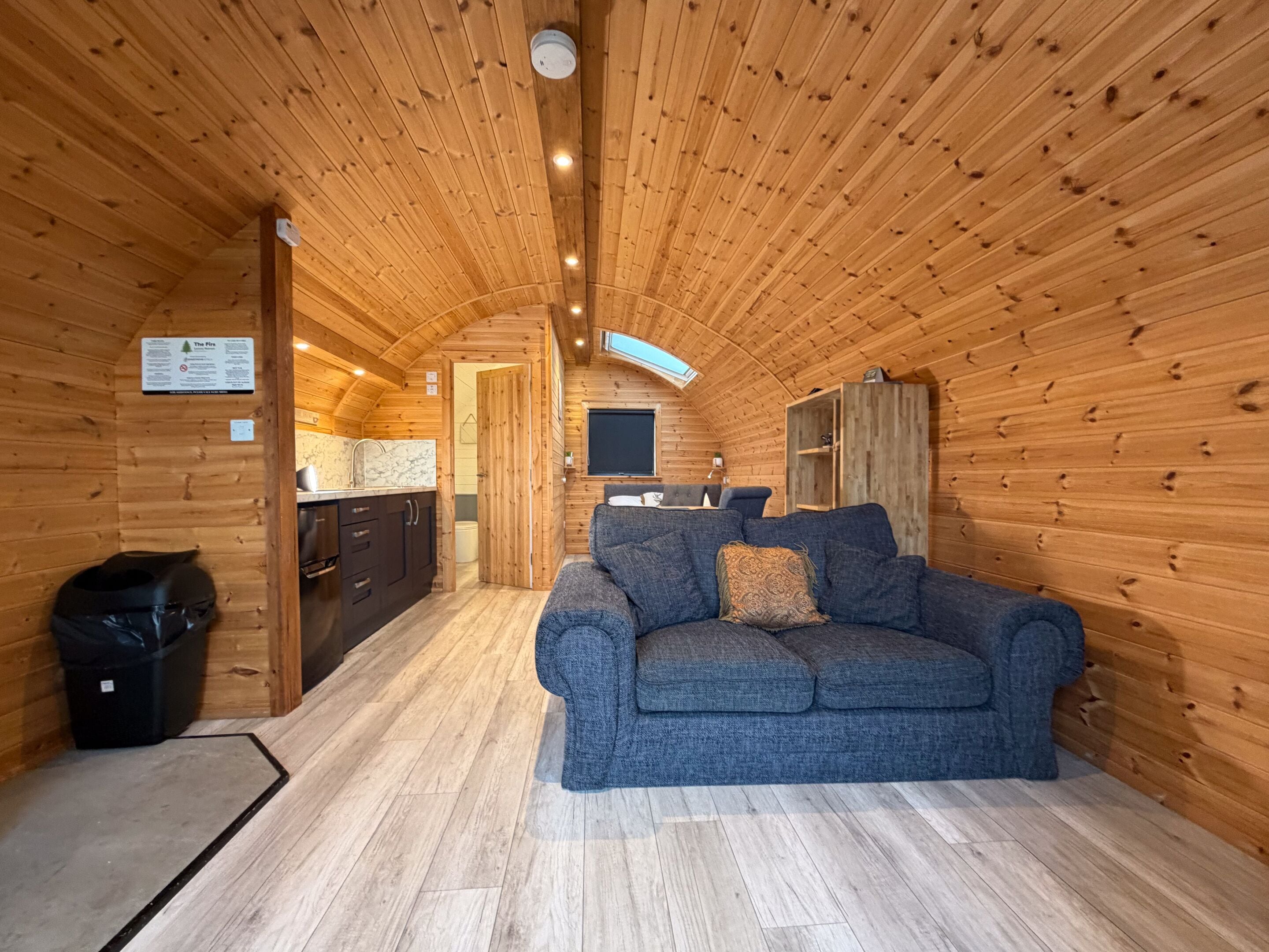 A cozy cabin interior with a wooden ceiling, a sofa, a kitchen area, and a fireplace.
