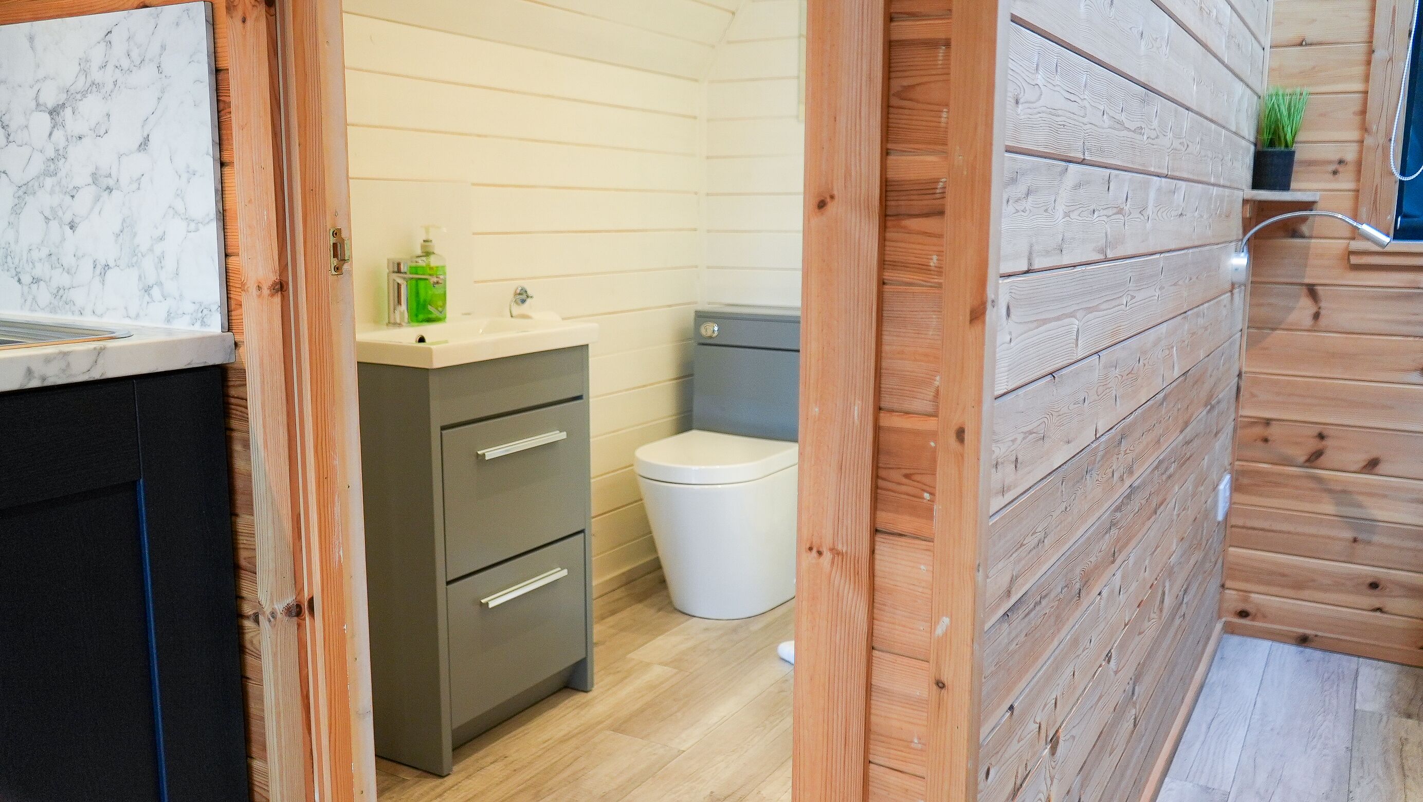 Une petite salle de bain avec un mur en bois, des toilettes blanches et un petit lavabo sur lequel est posée une bouteille verte.
