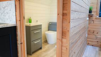 Une petite salle de bain avec un mur en bois, des toilettes blanches et un petit lavabo sur lequel est posée une bouteille verte.