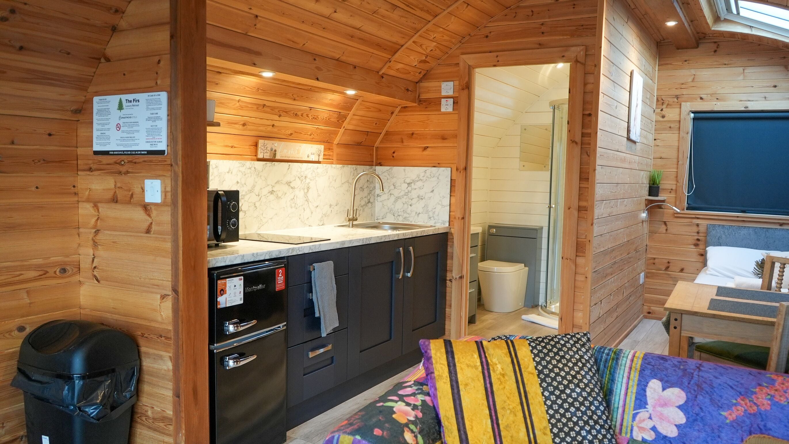 A compact, wooden cabin-style room with a kitchenette, a shower area, and a bed with colorful bedding.