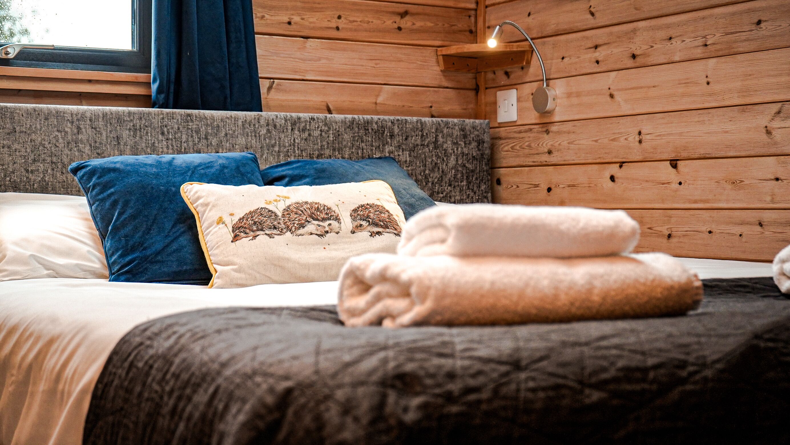 A cozy bedroom with a wooden wall, a bed with blue and beige pillows, and a bedside lamp.