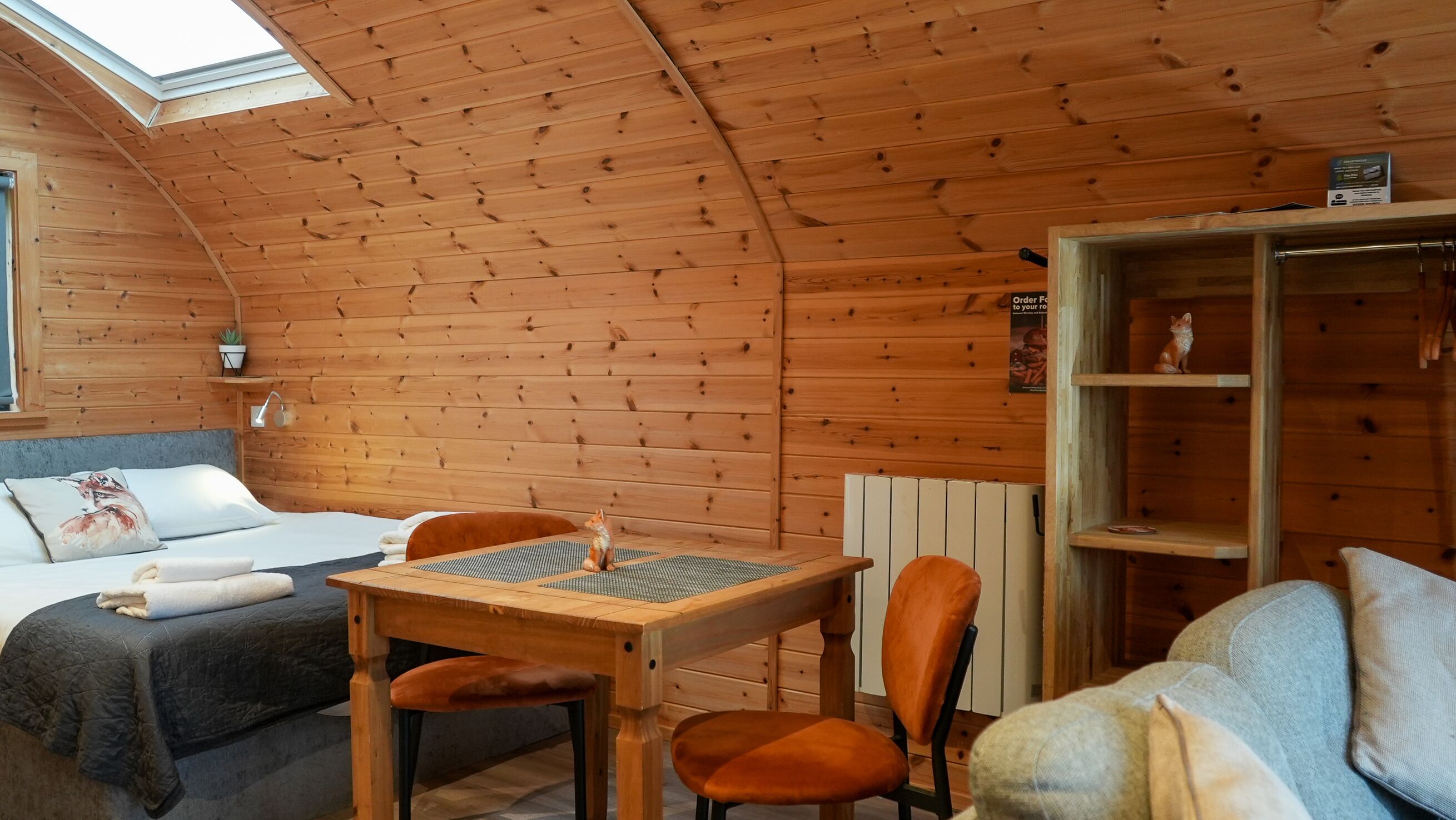 A cozy cabin room with a wooden ceiling, a bed with a pillow and a blanket, a small table with a chair, and a shelf with a figurine.