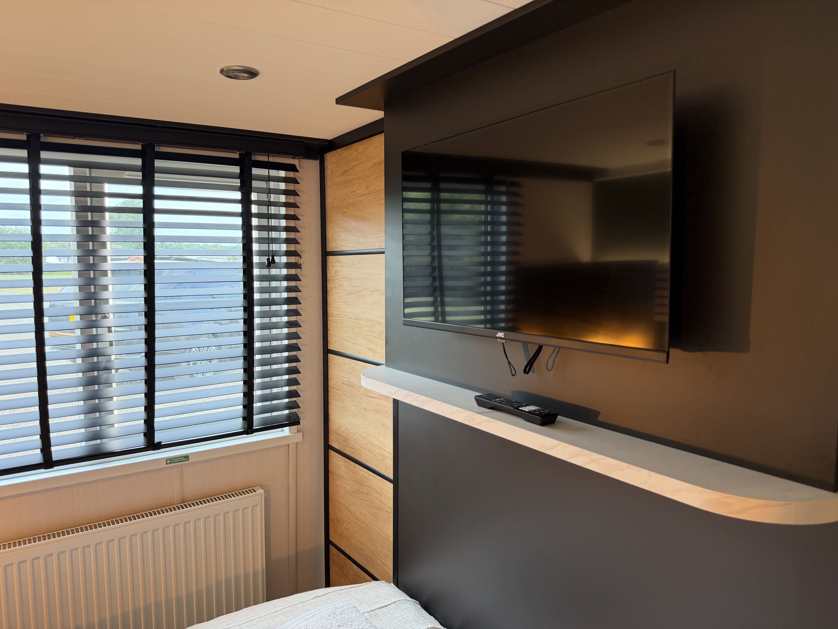 A modern room with a large flat-screen TV mounted on a wall, a window with blinds, and a built-in heater.