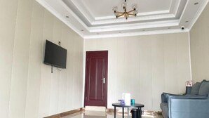 A spacious living room with a flat-screen TV mounted on the wall, a dark red door, a blue sofa, and a small round table with a lamp. - Sanmen Longyoujia Boutique Hotel (Sanmen)