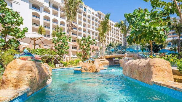 Zona de piscina de hotel con agua azul cristalina, formaciones rocosas, palmeras y un edificio de varios pisos al fondo.