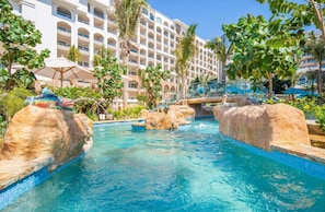 A hotel pool area with clear blue water, rock formations, palm trees, and a multi-story building in the background. - Valencia located between Tuly Beach and tequila Cove  (San José del Cabo)