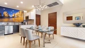 Cocina moderna con comedor, que incluye una mesa de vidrio, sillas blancas y un cuadro grande con una playa.