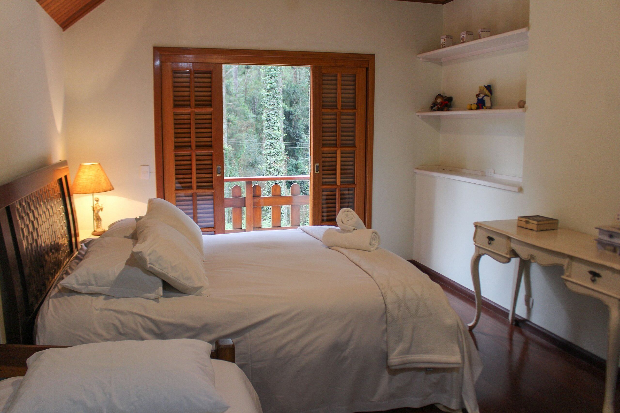 A bedroom with a bed, a nightstand, a lamp, a desk, and a window with shutters.