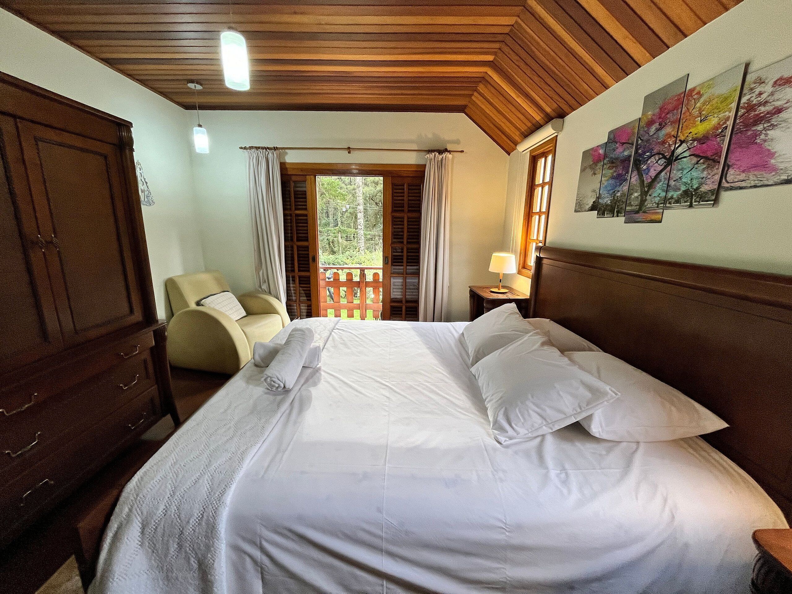 Een slaapkamer met een houten plafond, een bed met witte lakens, een stoel, een nachtkastje met een lamp en een raam met gordijnen.