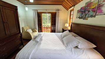 Een slaapkamer met een houten plafond, een bed met witte lakens, een stoel, een nachtkastje met een lamp en een raam met gordijnen.