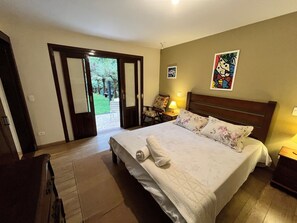 Un dormitorio con cama, mesita de noche, una silla, una puerta que da a un jardín y un muro con dos cuadros.