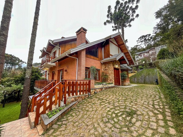 A two-story house with a stone facade, wooden balcony, and a paved driveway. - Casa Horto Village (Campos do Jordão)
