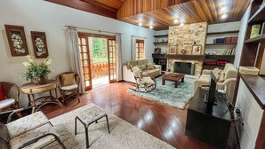 A spacious living room with a fireplace, wooden ceiling, and a view through a glass door. - Casa Horto Village (Campos do Jordão)