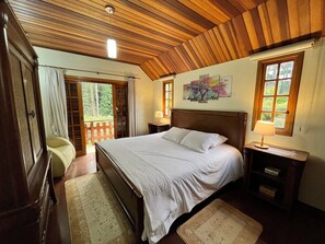 Un dormitorio con techo de madera, una cama con ropa blanca, una mesita de noche con una lámpara y una ventana con vistas a zonas verdes.