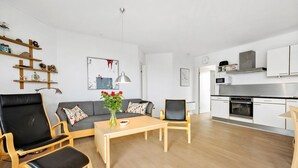 Ein modernes Wohnzimmer mit einer Couch, zwei Sessel, einem Couchtisch und einer Blumenvase. Es gibt eine Küchenzeile mit Herd und Schränken.
