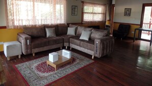 A living room with a brown sectional sofa, a small white ottoman, and a glass-top coffee table. There is a patterned rug on the wooden floor, and the room has wooden beams on the ceiling.