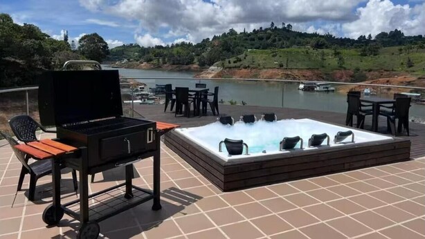 An outdoor patio with a grill, hot tub, and seating area overlooking a body of water and hills.