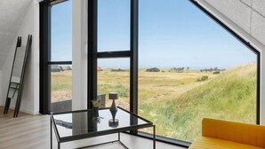 Una habitación moderna con ventanas amplias, una mesa de vidrio, un sofá amarillo y una escalera apoyada en la pared.