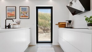 Una cocina moderna con armarios blancos, un grifo negro y una cafetera. Hay dos cuadros en la pared y una vista de un paisaje desértico a través de la puerta.