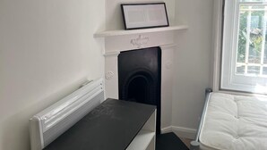 A small, clean room with a black table, a white bed, a fireplace, and a window with a view of greenery.