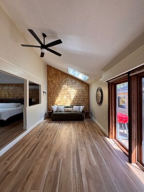 Una habitación amplia con suelo de madera, ventilador de techo, banco empotrado y un gran ventanal.