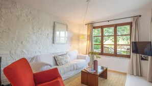 A living room with a white textured wall, a white sofa with patterned cushions, a red armchair, a wooden coffee table with a book and a vase, a TV on a stand, and a window with a view of greenery. - Flor de Pita Jimena de la Frontera by Ruralidays (Cádiz)