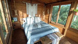 Une chambre à coucher dans une cabane en bois, avec un lit, des tables de chevet, une décoration en macramé suspendue et une fenêtre donnant sur les arbres.