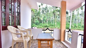 A covered patio with a wooden table and plastic chairs, overlooking a lush garden with tall trees. - Madhuvana Homestay Coorg  (Madikeri)