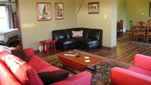 A living room with a red sofa, a black leather couch, and a wooden coffee table. There are framed pictures on the wall and a dining area in the background. - The Farm House - Quiet Country Retreat! Great for Any Season - Pet Friendly! (Osborn)