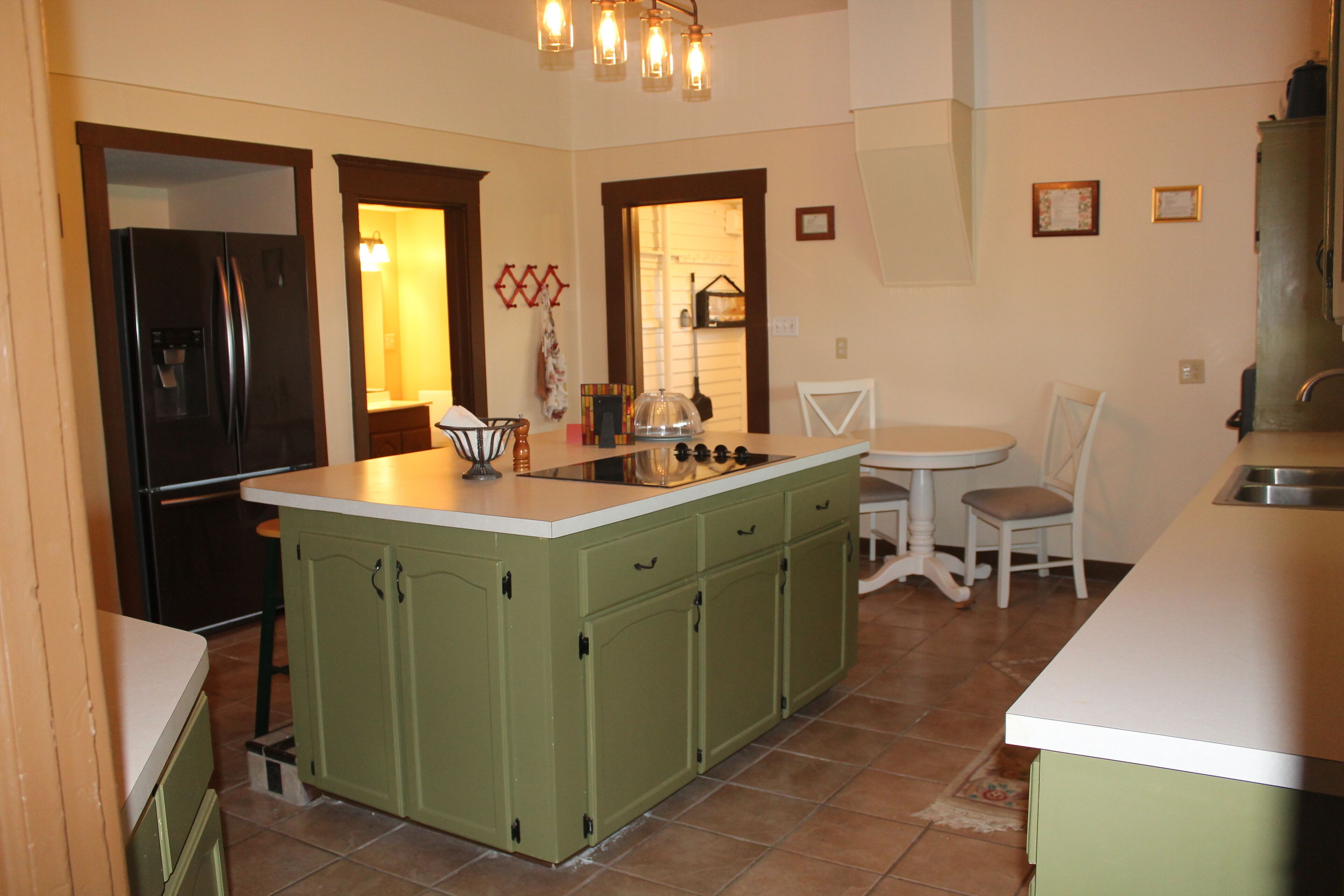 A kitchen with green cabinets, a black refrigerator, a white countertop, and a dining area with a white table and chairs.