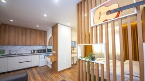 Ein kompaktes Hotelzimmer mit Küchenzeile, einem Bett mit grüner Bettdecke, einer Gitarre an der Wand und einer Pflanze auf dem Nachttisch.
