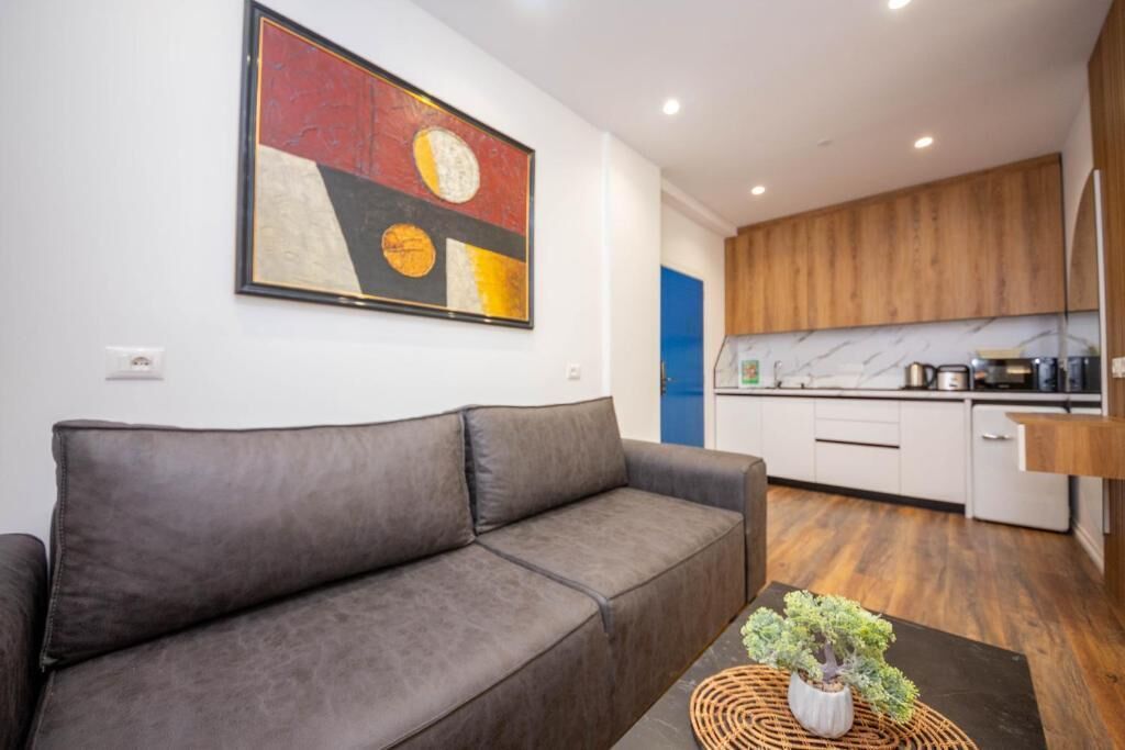 A modern living room with a grey sofa, a coffee table with a plant, and a kitchen in the background with wooden cabinets and white appliances.