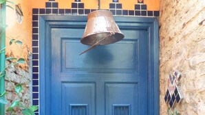 A blue door with a metal bell above it, set in a narrow alleyway with a plant on the left and decorative tiles on the right.