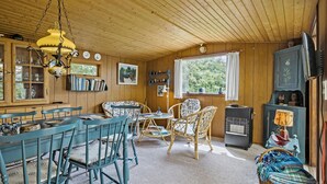 Ein gemütliches Blockhaus-Interieur mit Essbereich, einem Wohnzimmer mit Rattanmöbeln, einem Heizgerät und einem Fenster mit Blick auf Grünflächen.