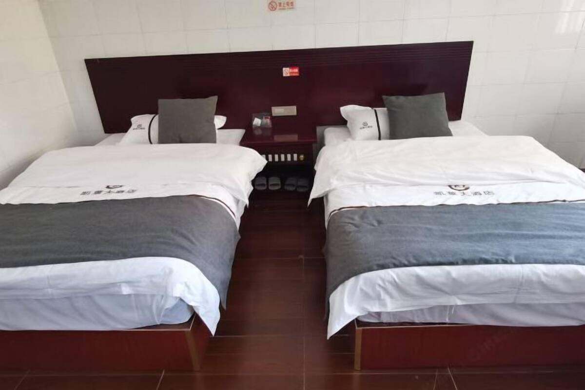Habitación de hotel con dos camas, cada una con ropa de cama blanca y gris, un cabecero oscuro y una mesita de noche pequeña con una lámpara.