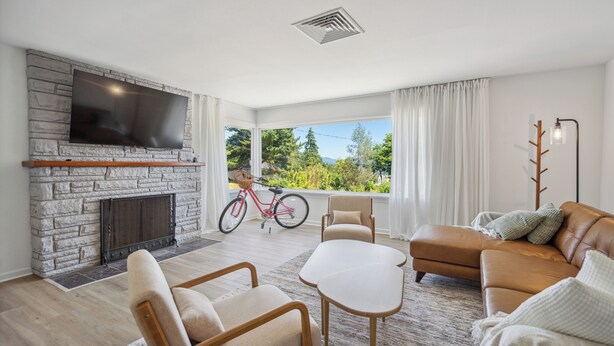 A modern living room with a stone fireplace, a flat-screen TV, comfortable seating, and a large window with a bicycle in the background. - Spacious 5B2B home 5 min to airport with AC, Game Room, Sleeps 16 (SeaTac)