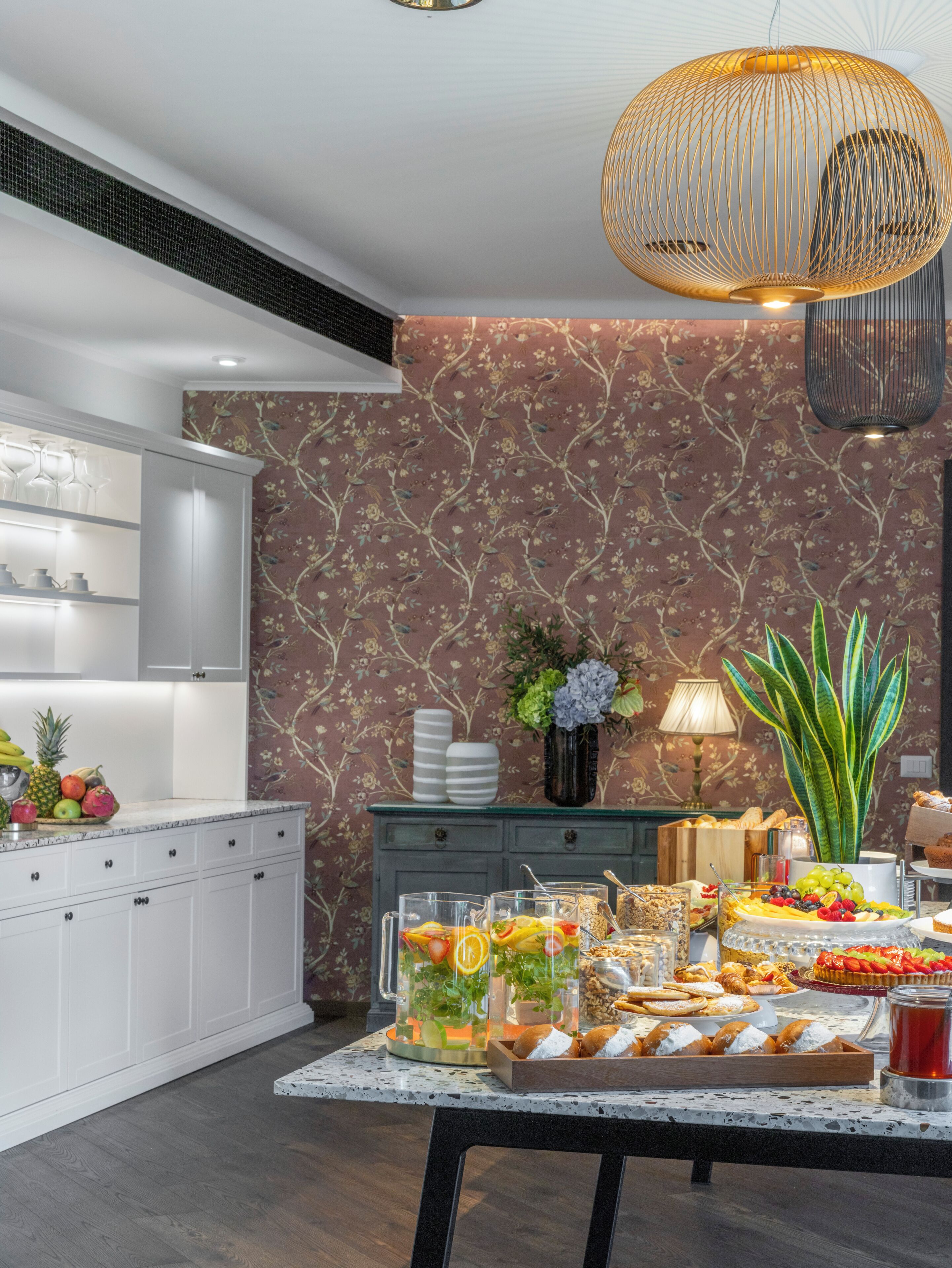 A well-lit room with a dining table set for a buffet, a decorative hanging light fixture, and a floral wallpaper pattern.