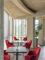 A modern interior with a conference table surrounded by red chairs, large windows with vertical blinds, and a potted plant.