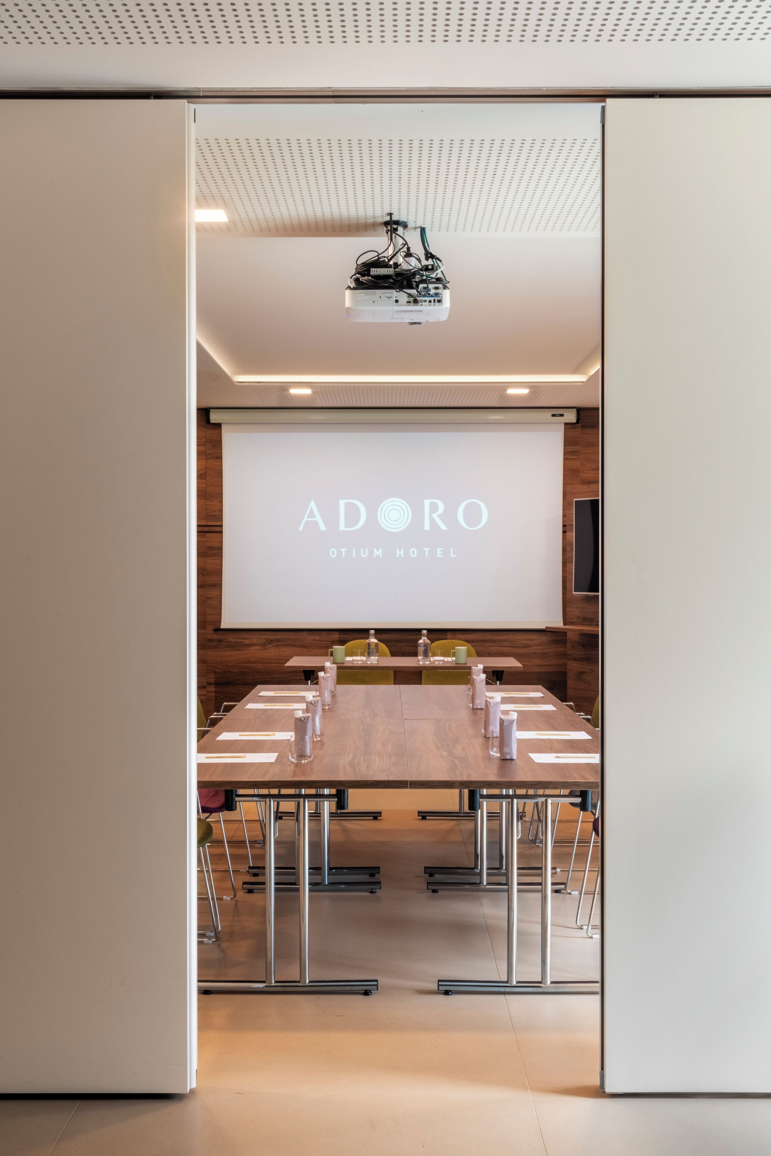 A conference room with a large projection screen displaying 'ADORO OTIUM HOTEL' branding, a long table with chairs, and a ceiling-mounted projector.