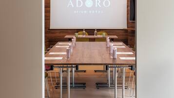 A conference room with a large projection screen displaying 'ADORO OTIUM HOTEL' branding, a long table with chairs, and a ceiling-mounted projector.