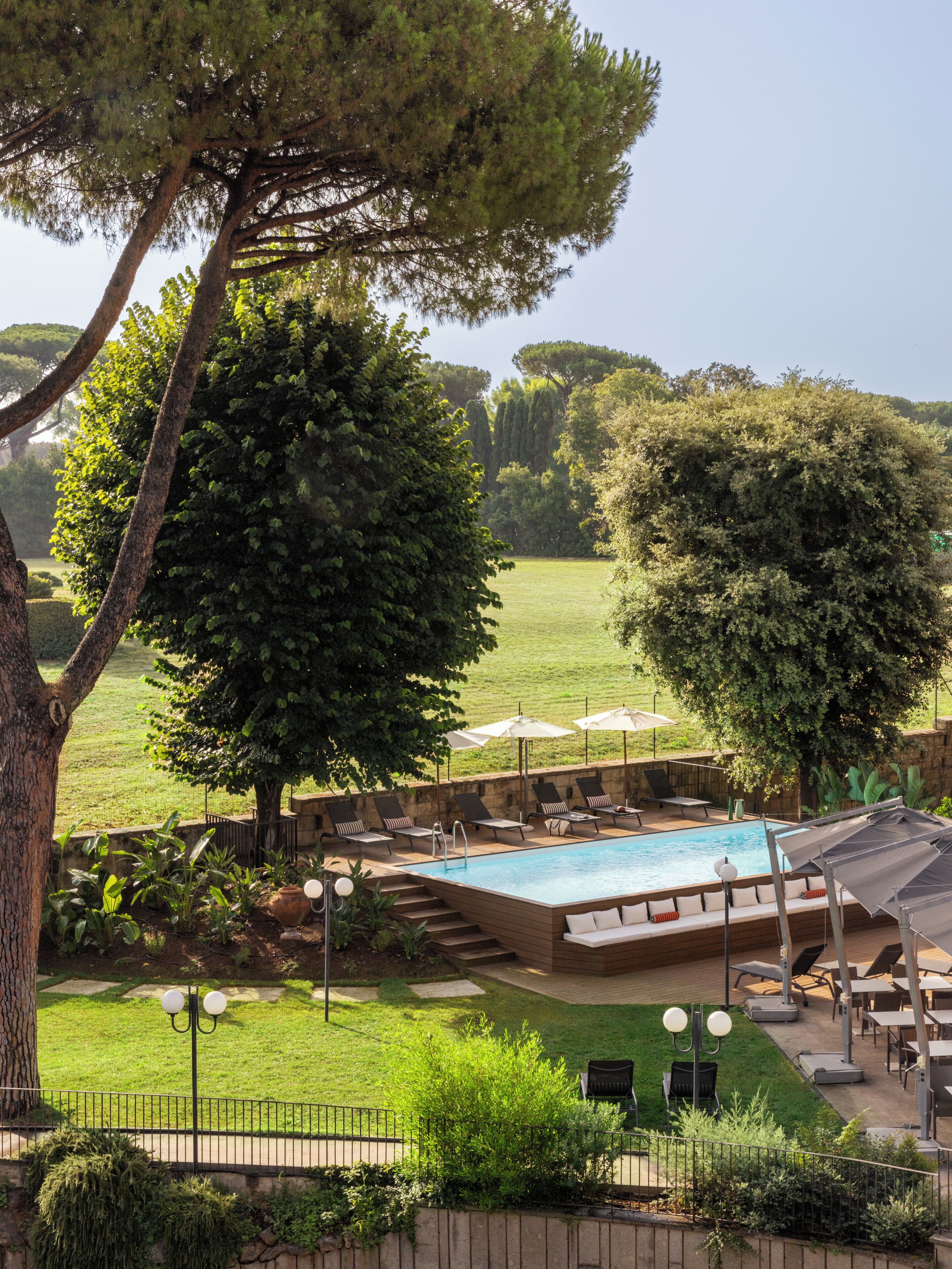 Un espace piscine doté de chaises longues et de parasols, entouré d’arbres et d’une pelouse verdoyante.