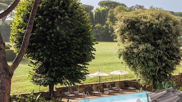 Un espace piscine doté de chaises longues et de parasols, entouré d’arbres et d’une pelouse verdoyante.